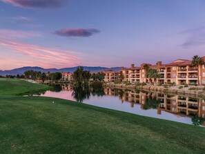 Un parcours de golf avec un lac paisible, dans lequel se reflètent des bâtiments dotés de balcons, et un ciel dégagé.