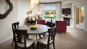 Quarto de hotel com área de refeições, conjunto de sofás, lareira, TV e vista para um campo de golfe.