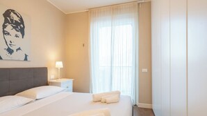 Hotelkamer met een groot bed, een nachtkastje met een lamp, een raam met dunne gordijnen en een kunstwerk aan de muur.