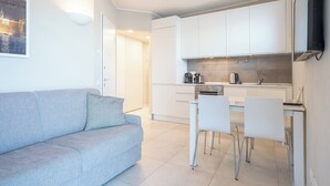 Een modern, licht appartement met een grijze bank, een eethoek met witte stoelen en een keuken met witte kastjes.