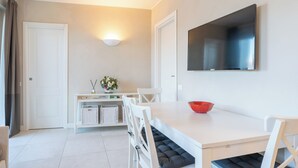 A modern dining area with a white table, chairs, and a red bowl. There is a TV mounted on the wall and a small shelf with a vase and candles.
