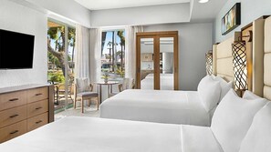 A hotel room with two beds, a TV, a desk with a chair, and a balcony with a view of palm trees.