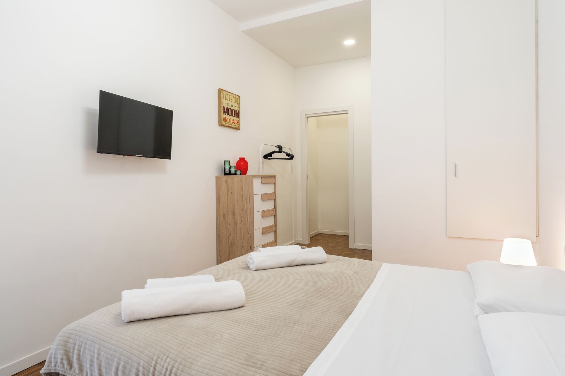 Ein Schlafzimmer mit einem Bett, einem an der Wand montierten Fernseher, einem Holzschrank und einer Tür zu einem anderen Raum.