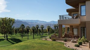 A modern building with a balcony overlooking a well-manicured lawn and palm trees, with mountains in the background.