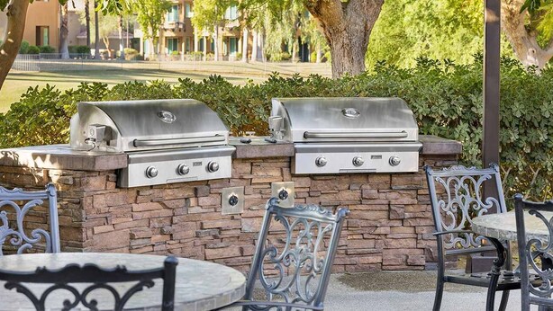 A patio with two stainless steel barbecue grills, stone walls, and wrought iron chairs.
