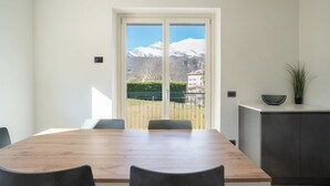 A modern dining area with a wooden table and chairs, a view of mountains through a glass door, and a small kitchen area with a plant.