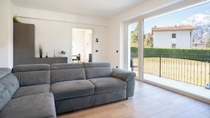 A modern living room with a grey sofa, a small dining area, and a balcony with a view of a mountain.
