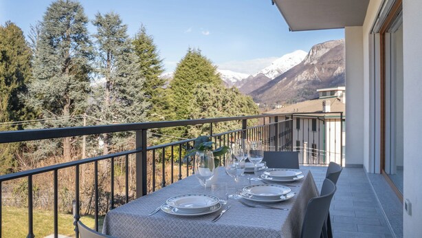 A balcony with a table set for dining, overlooking a mountainous landscape with trees and a building in the background.