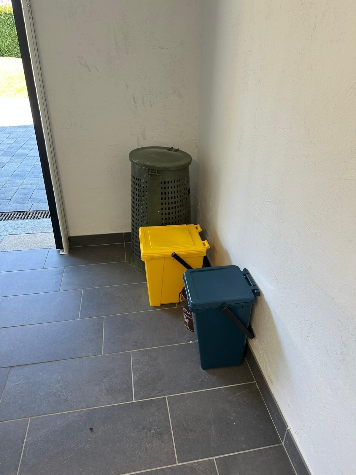 Eine Raumecke mit Fliesenboden, einem grünen Mülleimer und zwei Aufbewahrungsboxen, eine gelb und eine blau.