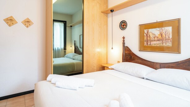 Una camera da letto con testiera in legno, un letto con lenzuola bianche, uno specchio, un quadro e un comodino con una lampada.