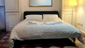 Siististi pedattu sänky, jossa on kuviollinen päiväpeite, sen yläpuolella oleva kehystetty peili ja yöpöydällä oleva lamppu. Huoneessa on ruutukuvioinen lattia ja valkoiset seinät, joissa on paneeli.