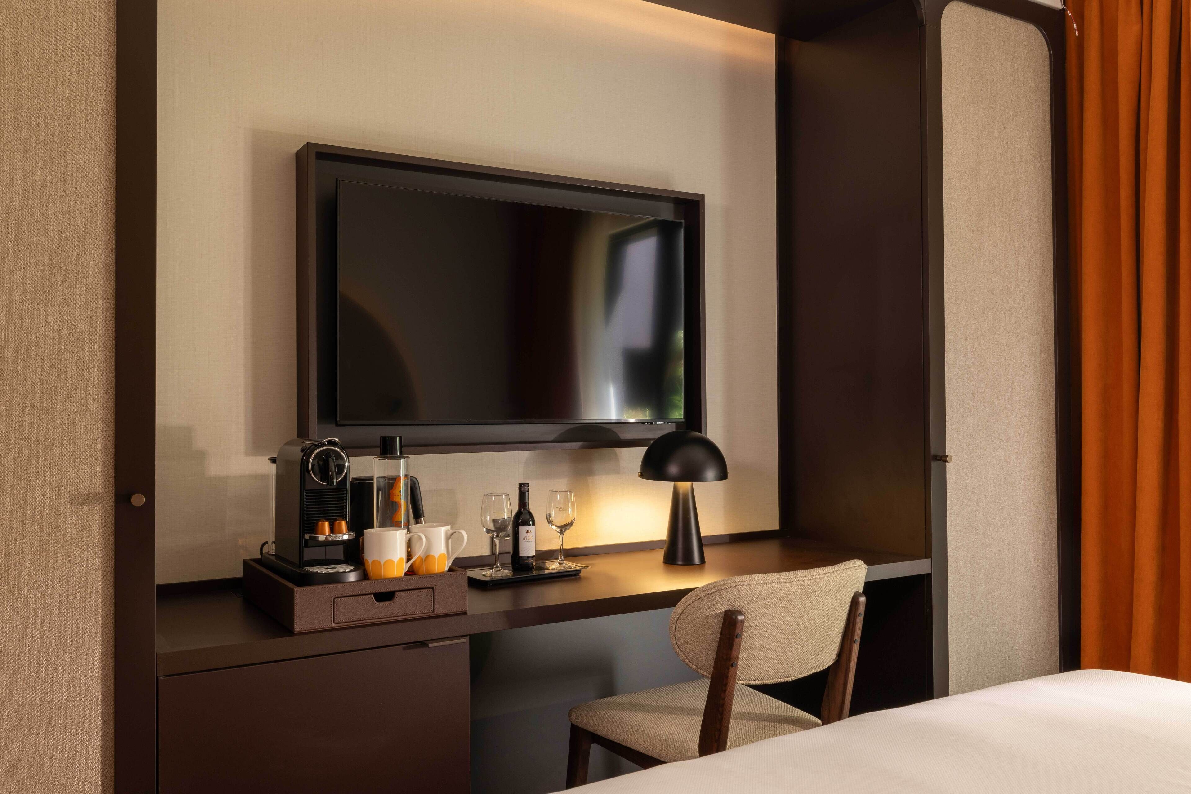 A hotel room with a flat-screen TV mounted on the wall, a desk with a coffee maker, glasses, and a wine bottle, and a bed with white sheets.