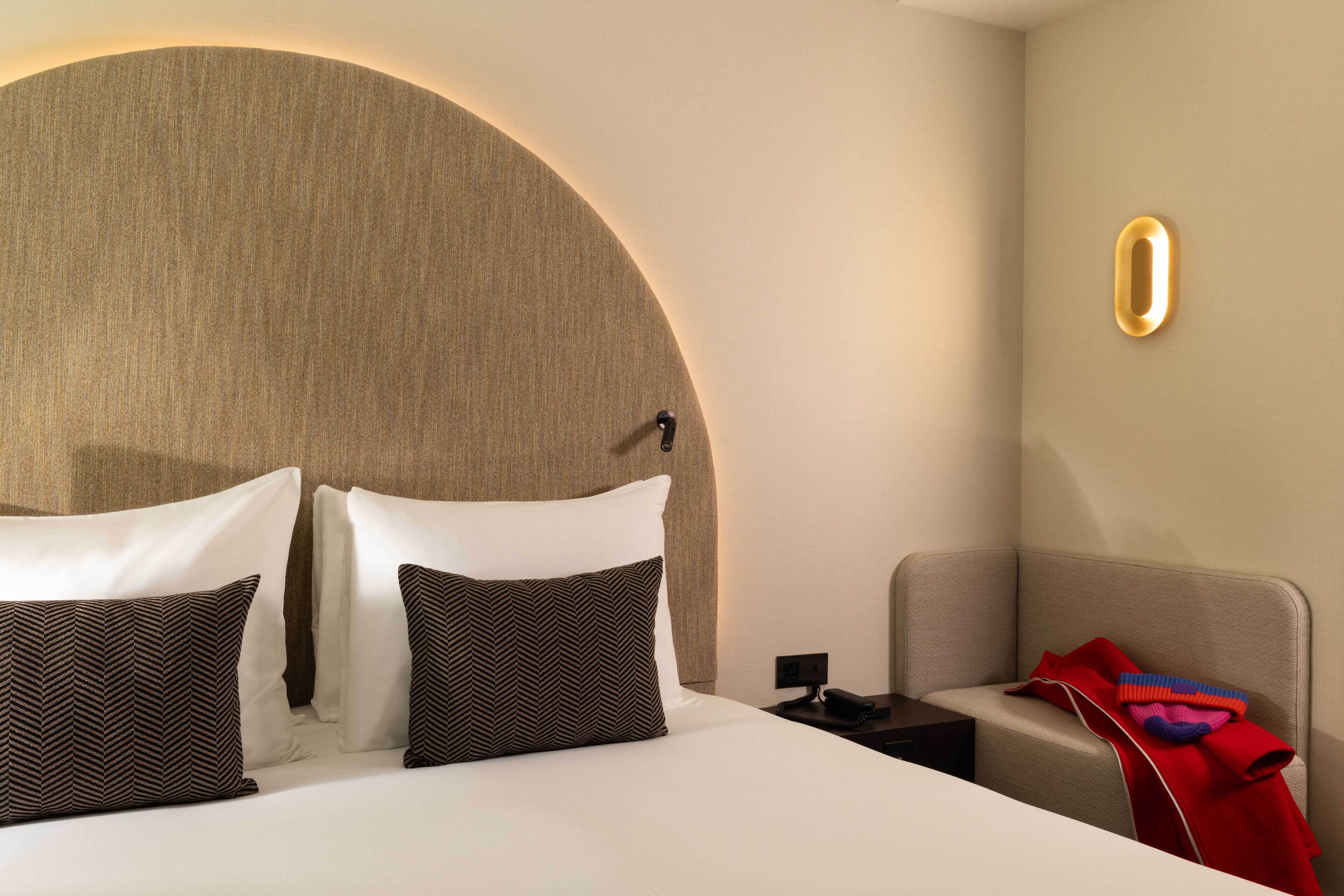 Ein modernes Hotelzimmer mit einem großen Bett, einem Kopfteil im Kreismuster, einem Nachttisch mit Telefon und einem roten Stoffüberwurf auf dem Sessel.