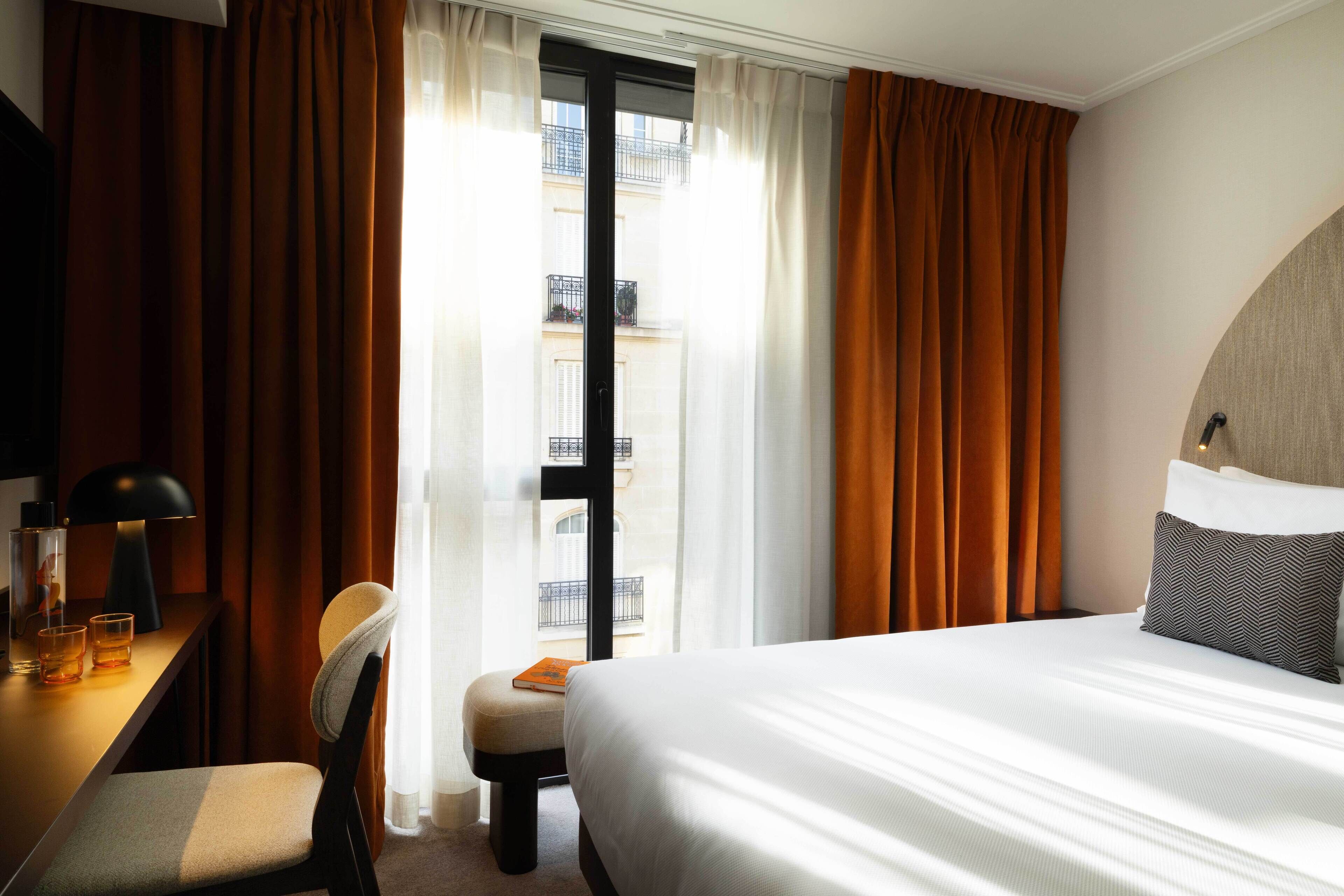 A hotel room with a bed, a desk with a lamp, a chair, and a view of a building through a window.