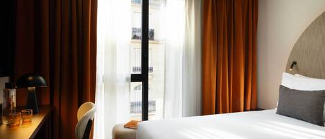 A hotel room with a bed, a desk with a lamp, a chair, and a view of a building through a window.