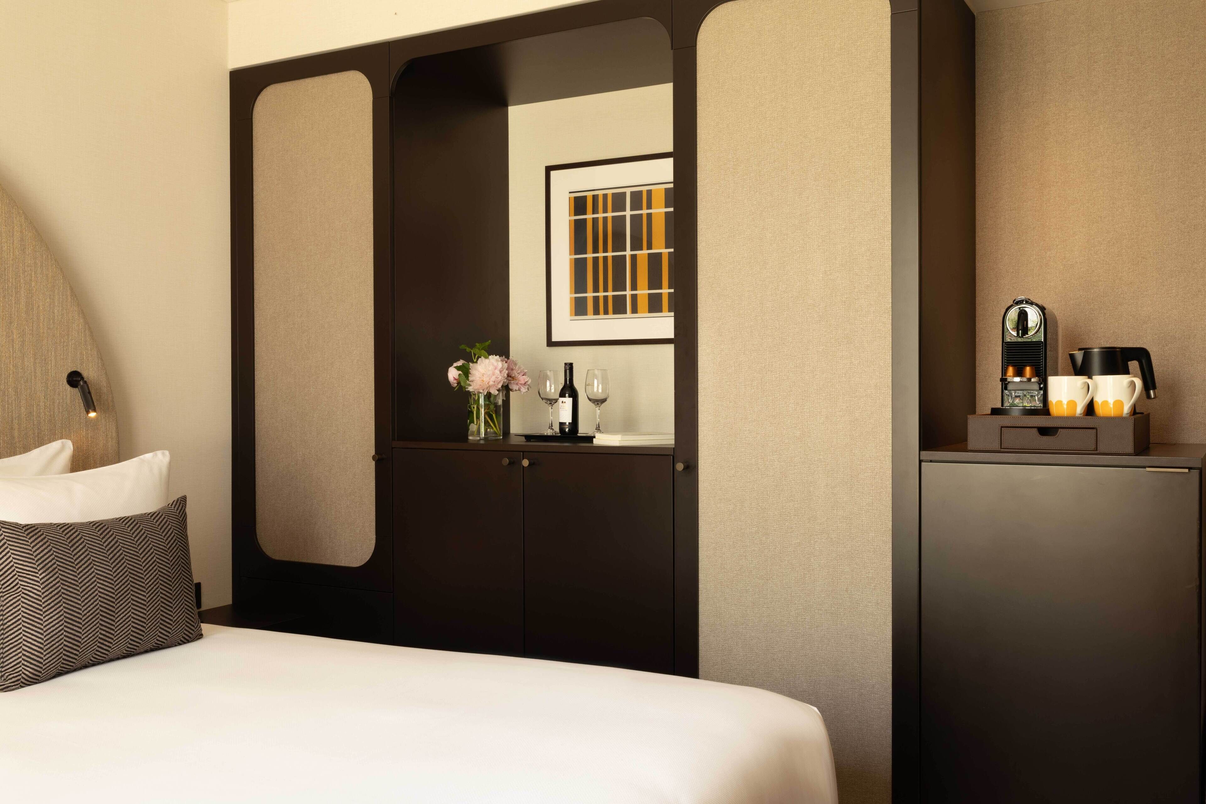 Una habitación de hotel con cama, cómoda, espejo, un cuadro enmarcado, cafetera y un florero con flores.