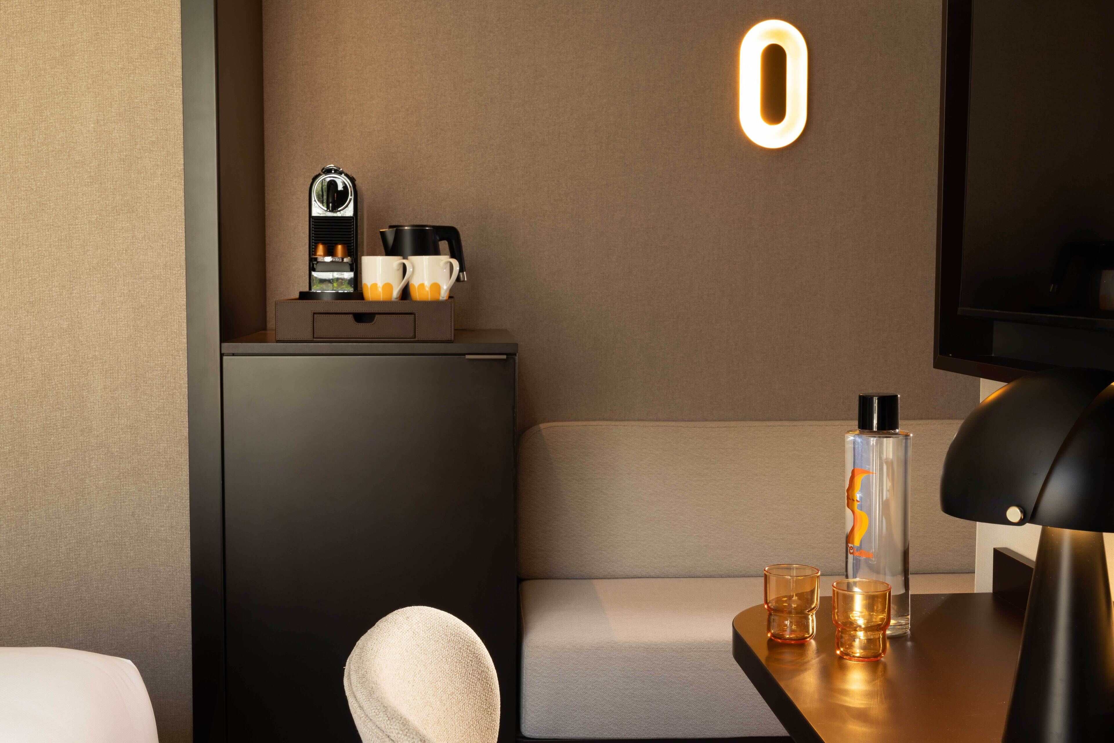 Una habitación de hotel moderna con sofá, una mesita con lámpara, minibar y televisión.