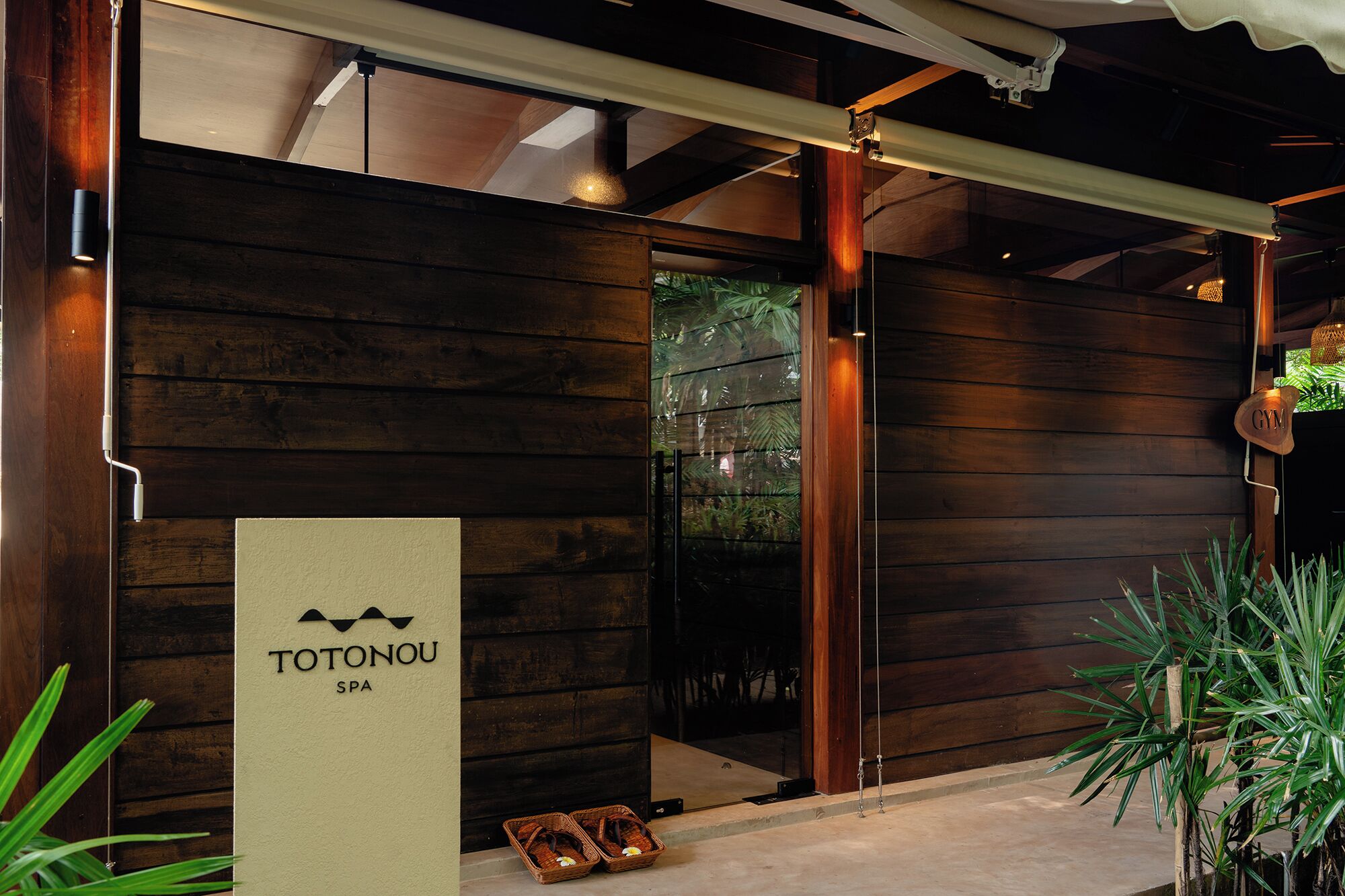 Fachada de madera de un edificio con el letrero 'TOTONOU SPA', puerta de vidrio de entrada, una lámpara colgante y una planta pequeña.