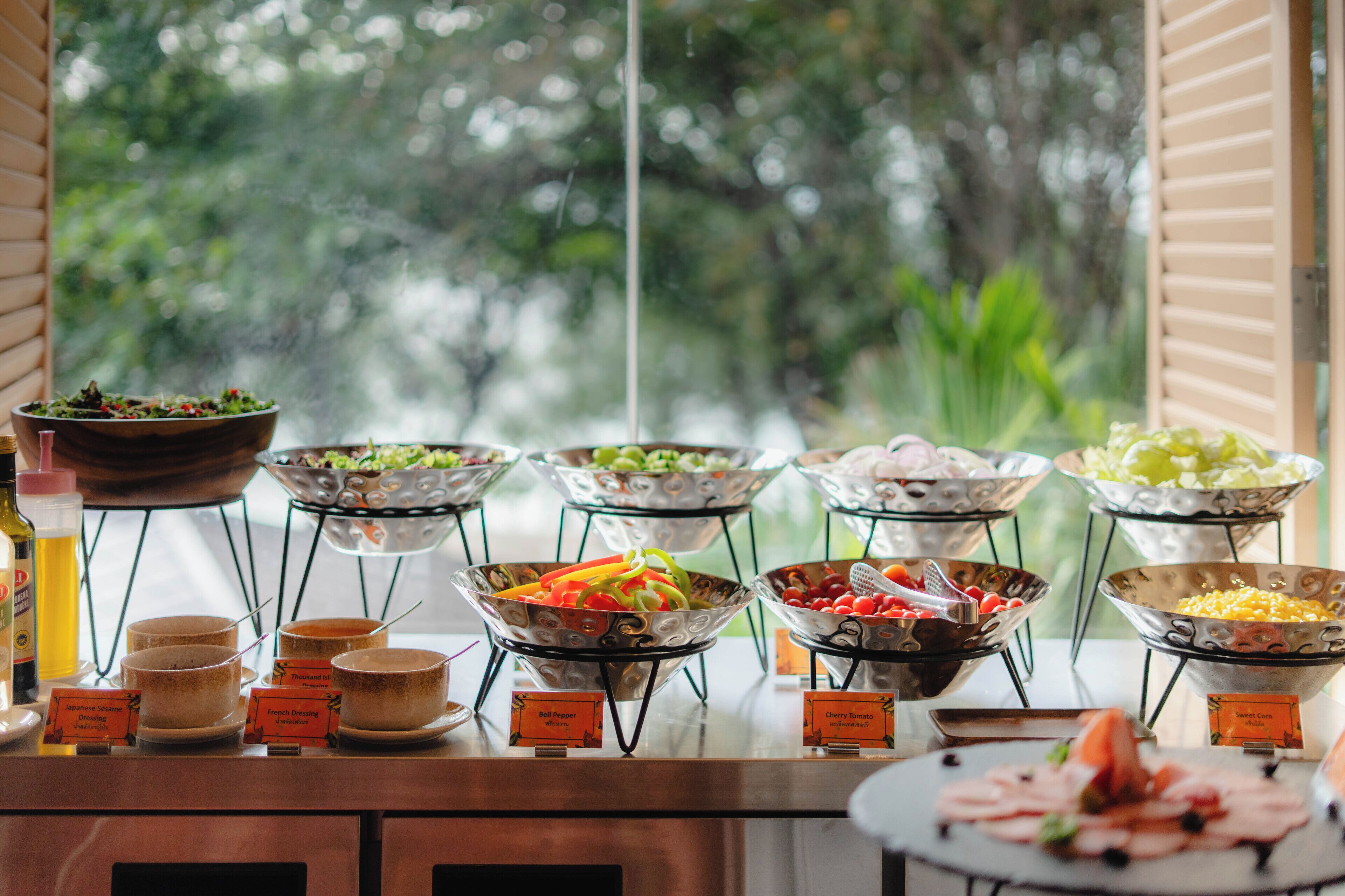 Un buffet proposant une variété de plats et d’aliments, chacun étant clairement identifié par une étiquette, le tout installé devant une fenêtre donnant sur un paysage verdoyant.