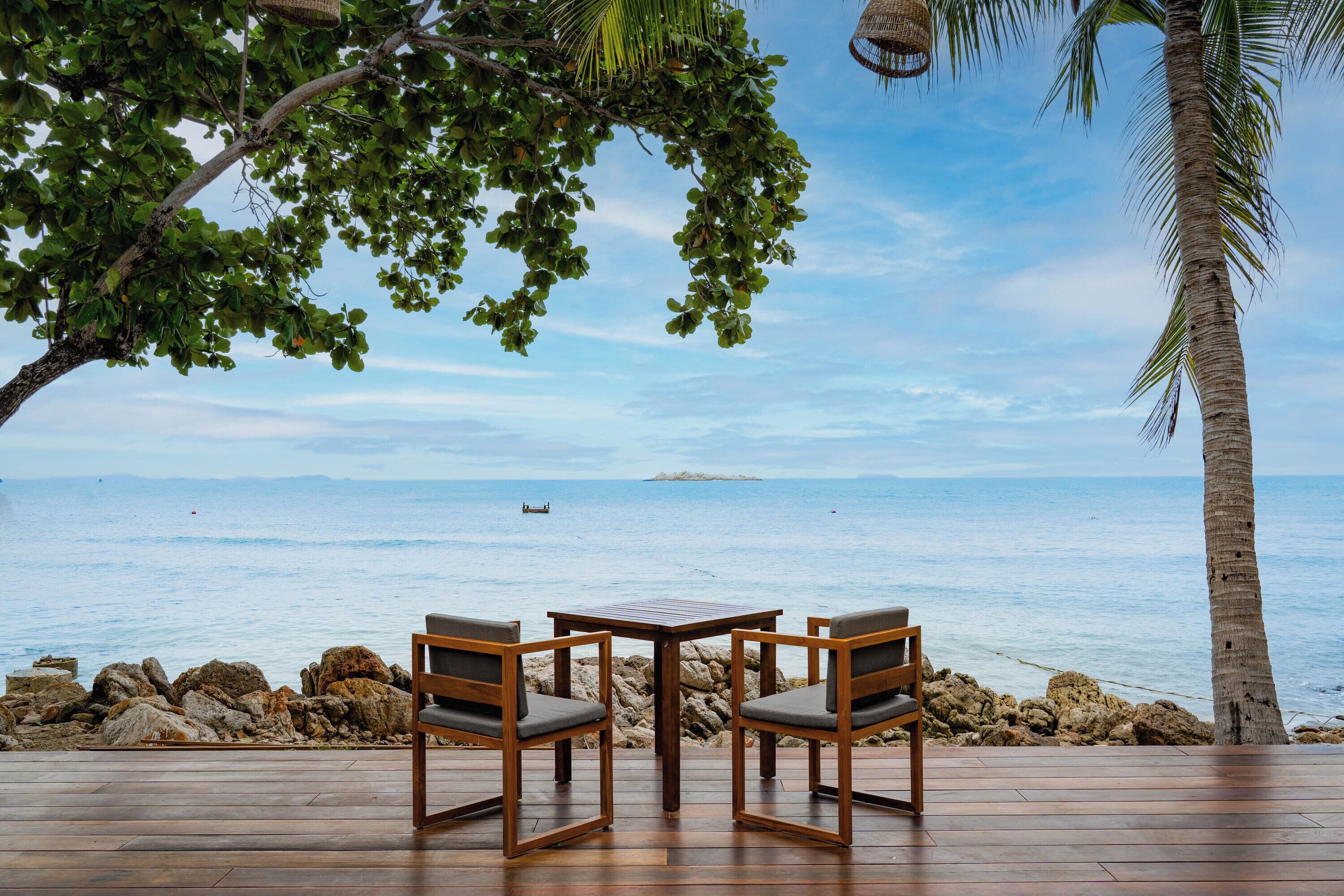 Une terrasse en bois avec deux chaises et une table, donnant sur un rivage rocheux et la mer.
