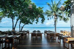 An outdoor dining area with wooden tables and chairs, overlooking a beach with palm trees and the ocean. - Kira Baya Koh Samed (Rayong)