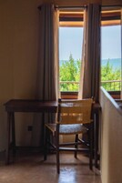 Una habitación con un escritorio y silla de madera, una ventana con cortinas, y vistas a follaje y al mar.