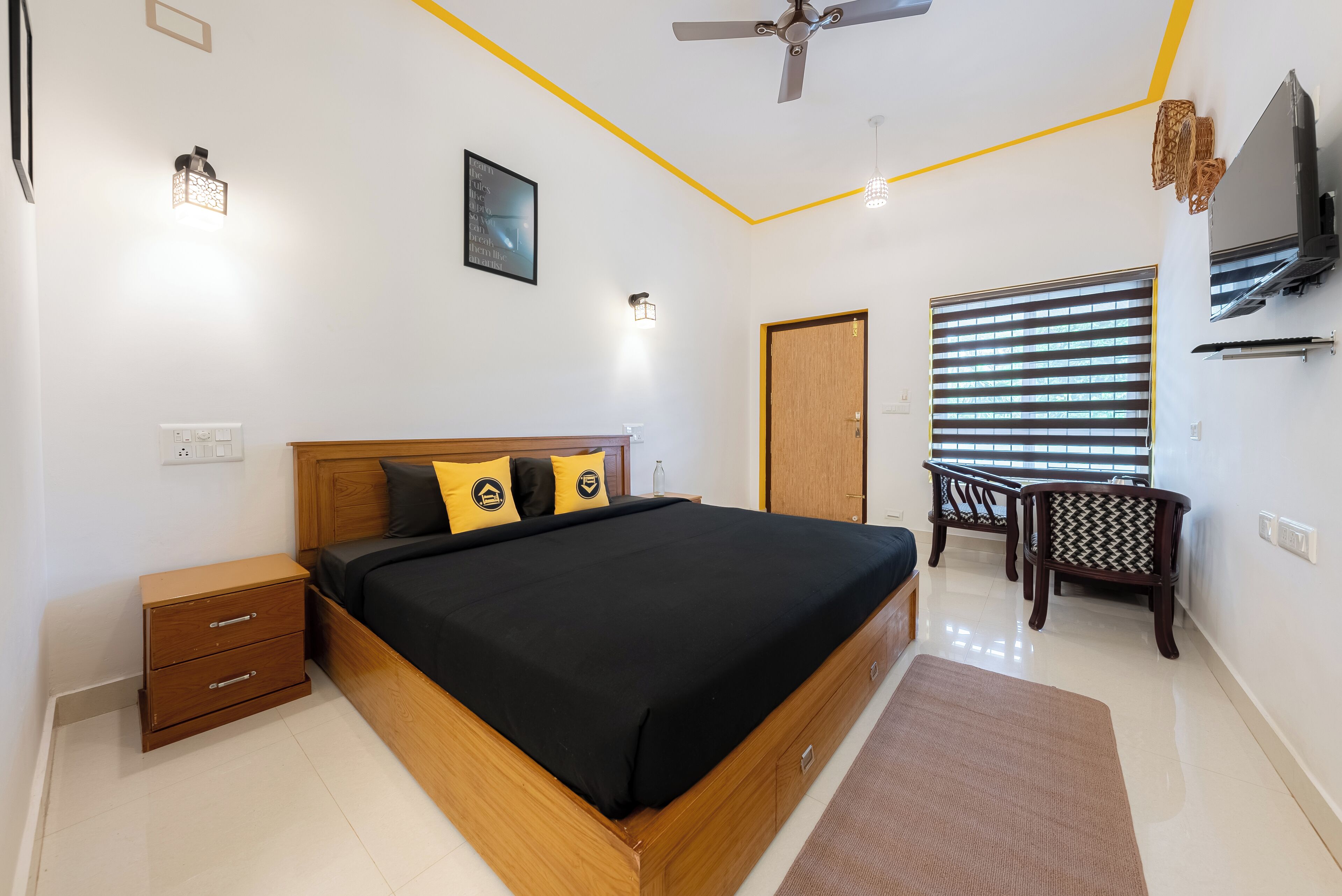 A bedroom with a wooden bed, two bedside tables, a ceiling fan, a window with blinds, and a TV mounted on the wall.