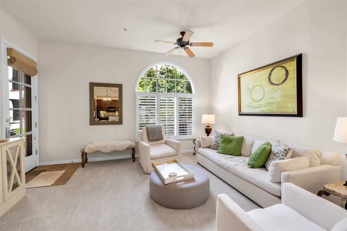 A spacious living room with a white sofa, a round ottoman, a ceiling fan, and a large window with shutters.