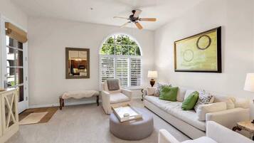 A spacious living room with a white sofa, a round ottoman, a ceiling fan, and a large window with shutters.