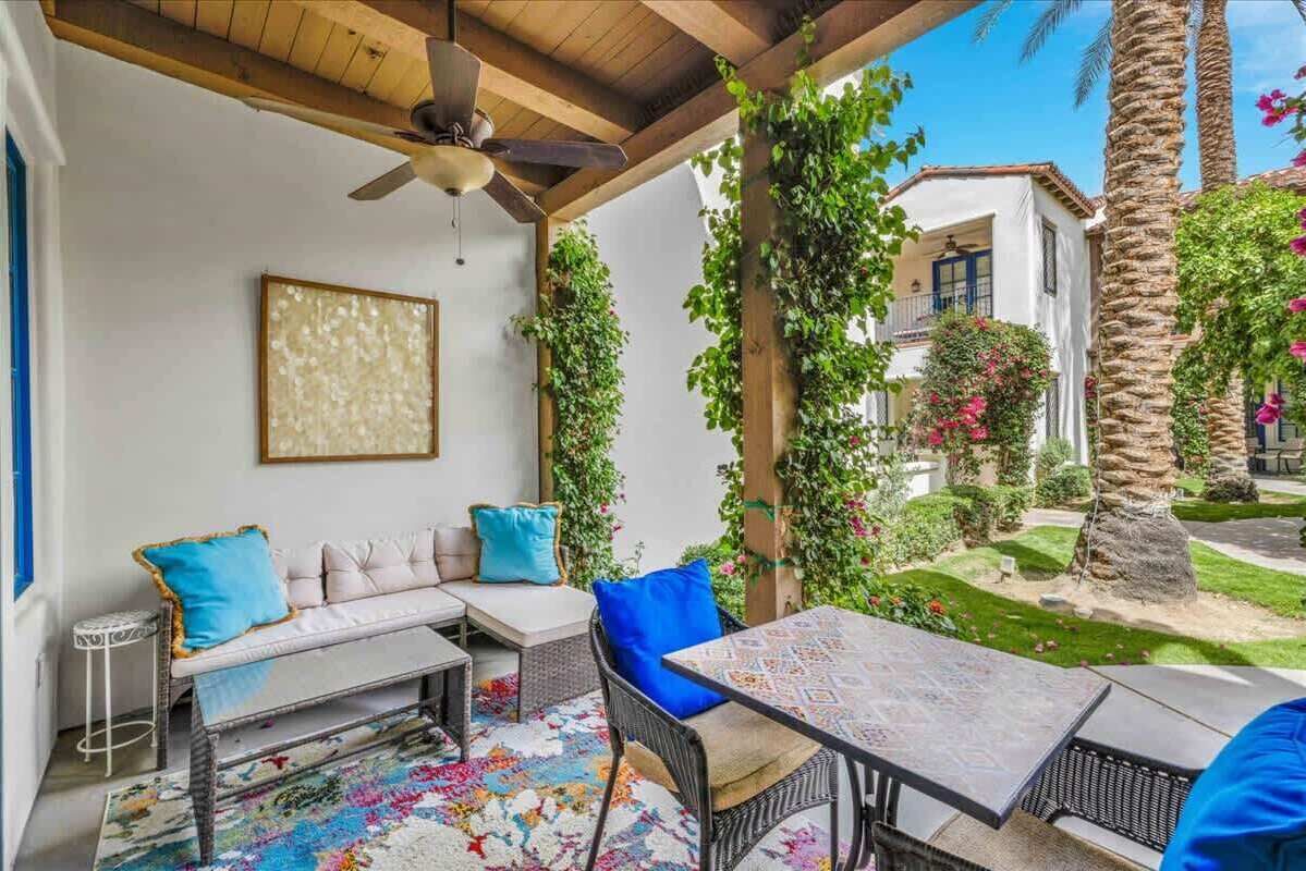 A covered outdoor seating area with a ceiling fan, a sofa with cushions, a table, and a patterned rug.