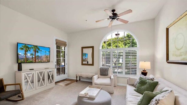 A spacious living room with a large TV, comfortable seating, a ceiling fan, and a view through a window with shutters. - Desert Charm A | 1BR Legacy Villa (La Quinta)