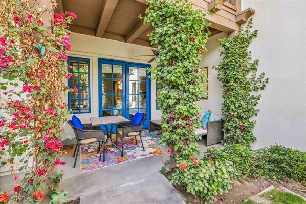 A covered patio with a table and chairs, a blue door, and a wall covered in climbing plants.