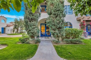 A house with a central archway, a paved pathway leading to a blue door, and a well-maintained garden with various shrubs and a small tree.