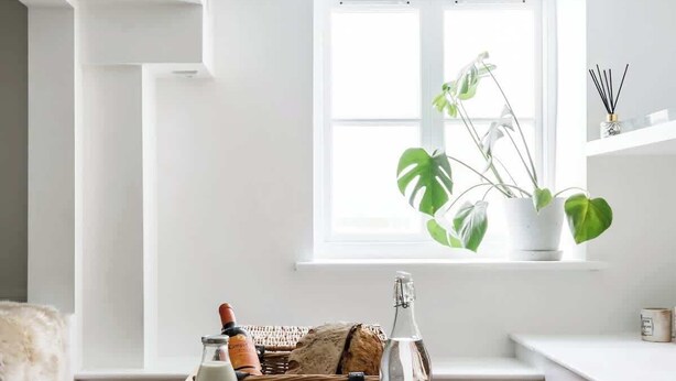 Ein heller, weißer Raum mit einem Holztisch und einer Bank, auf denen Gläser, eine Flasche und ein Korb stehen. Im Hintergrund ist ein Fenster mit Pflanzen zu sehen.