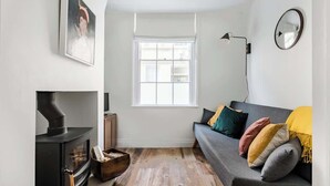 Ein modernes Wohnzimmer mit einem schwarzen Kaminofen, einem grauen Sofa mit bunten Kissen, einem großen Fenster und einem wandmontierten Spiegel.