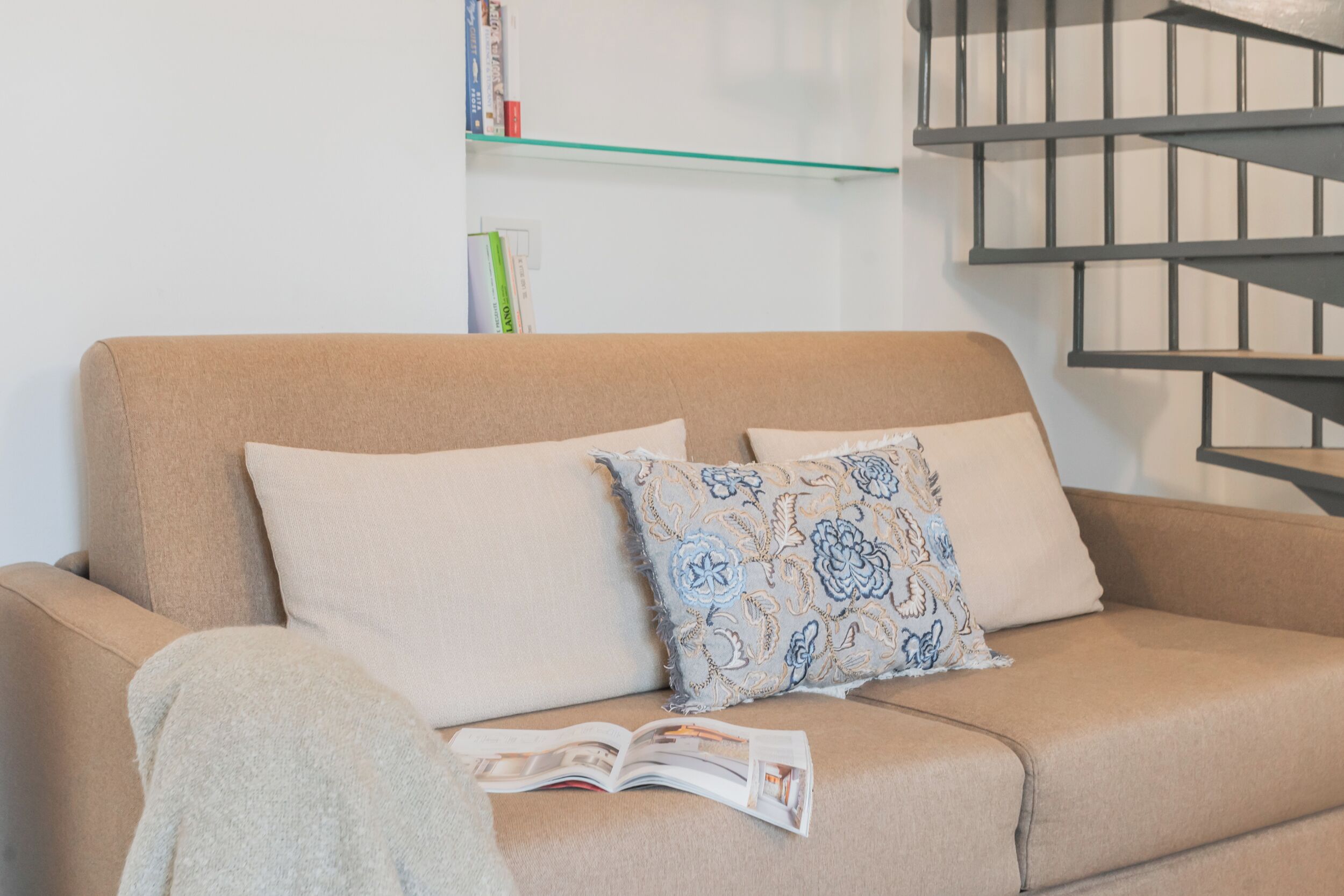 Ein beiges Sofa mit zwei Kissen, ein schlichtes und eines mit Blumenmuster, sowie eine Zeitschrift darauf.