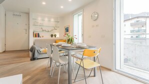 Un comedor moderno con una mesa de vidrio, sillas blancas y amarillas, y vistas a un balcón con edificios al fondo.
