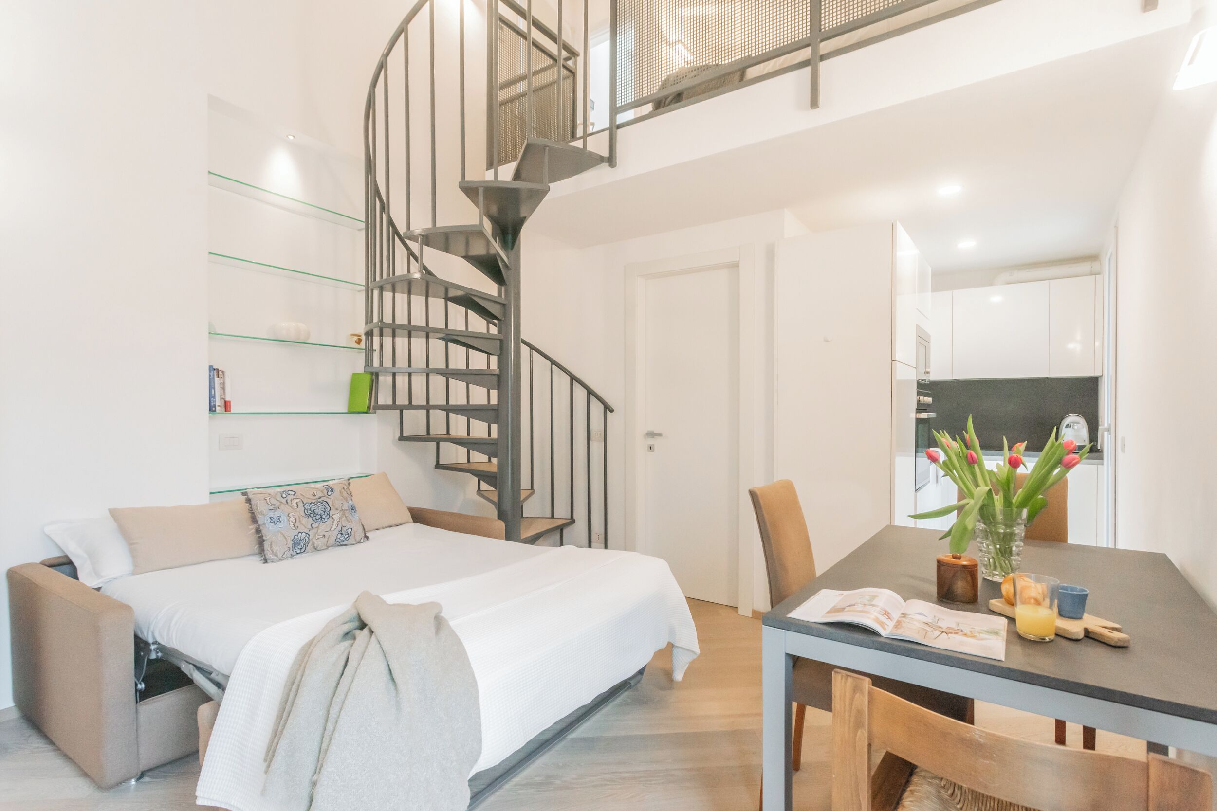 A modern apartment with a bed, a dining table, and a spiral staircase.