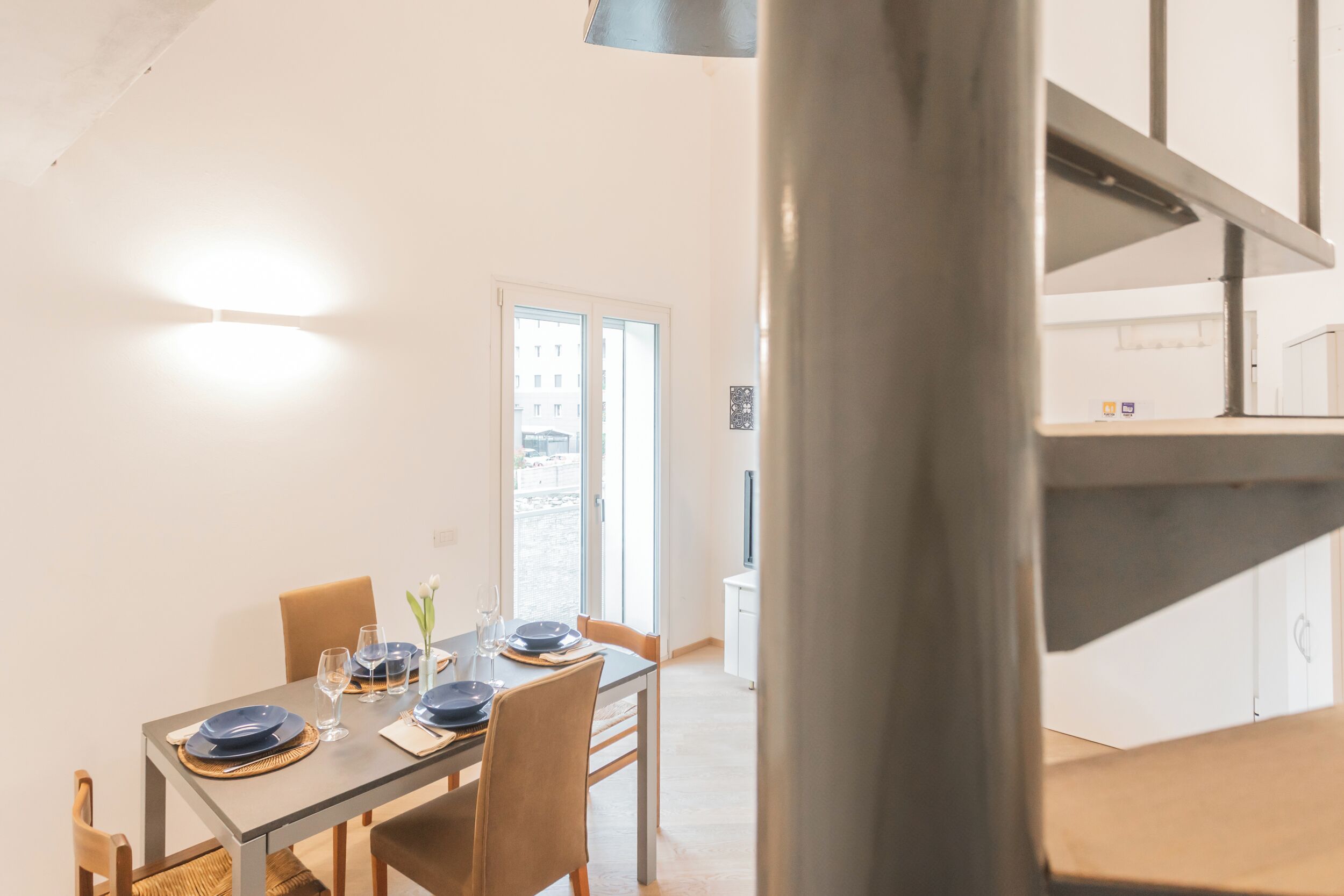 Un comedor con una mesa dispuesta para dos, vistas de la ciudad a través de un ventanal y una escalera que lleva a un nivel superior.