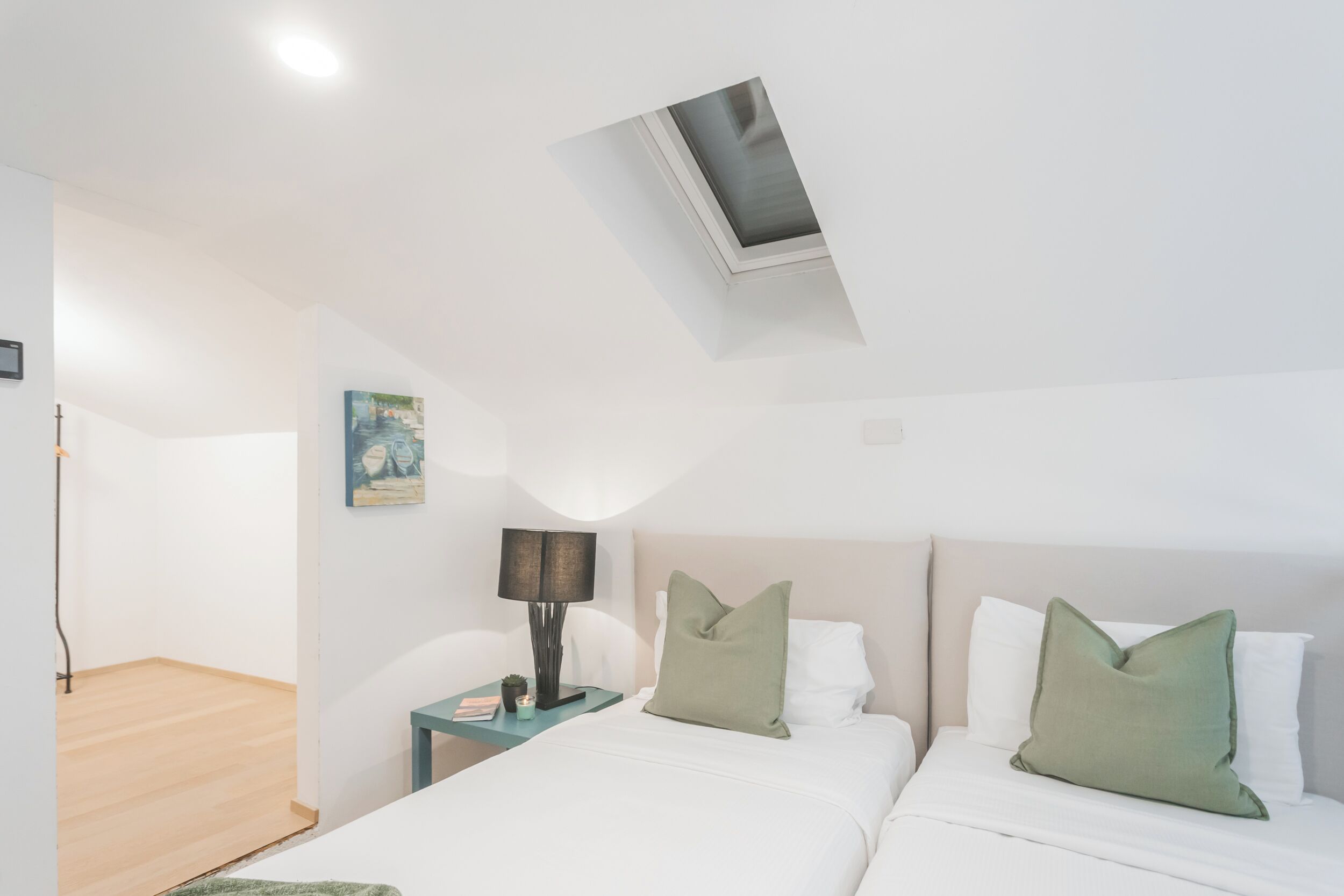 Un dormitorio con tragaluz, una cama con ropa de cama blanca y almohadas verdes, una mesita de noche con lámpara y una pequeña planta.
