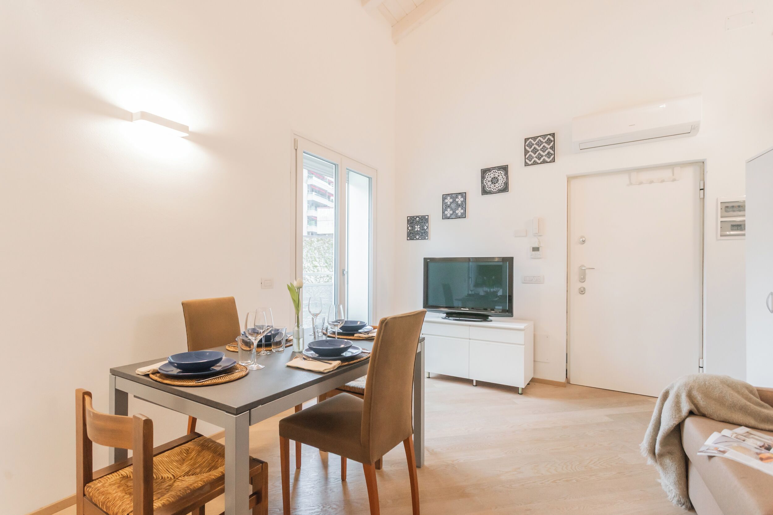 Un coin repas avec une table dressée pour quatre personnes, un téléviseur posé sur un meuble blanc et une porte équipée d’un appareil de climatisation.