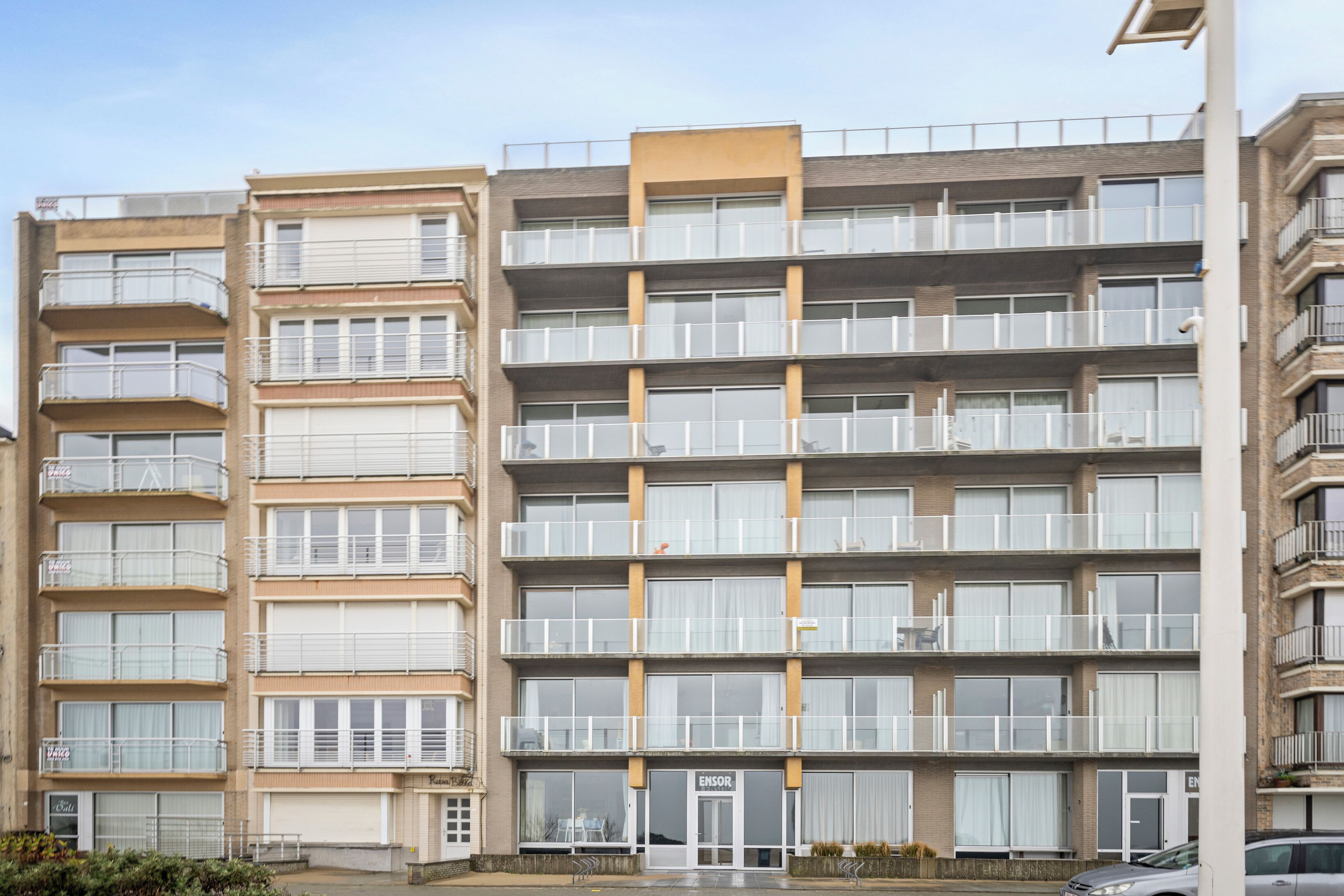 A multi-story residential building with balconies, a central entrance, and a sign reading "ENSOR".