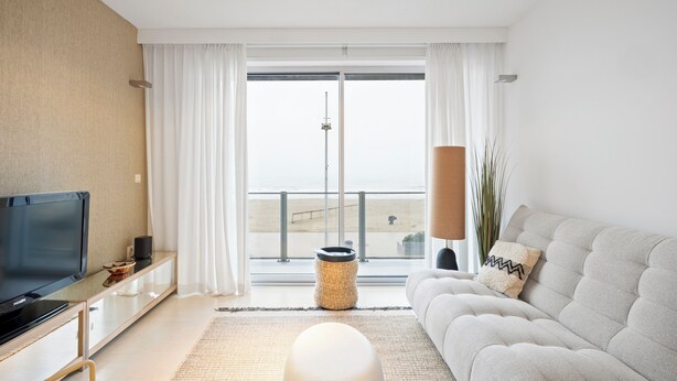 A modern living room with a television, sofa, and large windows overlooking a beach.