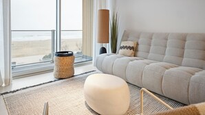 A modern living room with a large window, a beige sofa, a round ottoman, and a floor lamp.