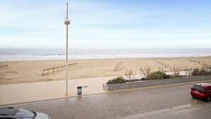 A beach view with a sandy shore, a tall pole, a red car, and a paved walkway.
