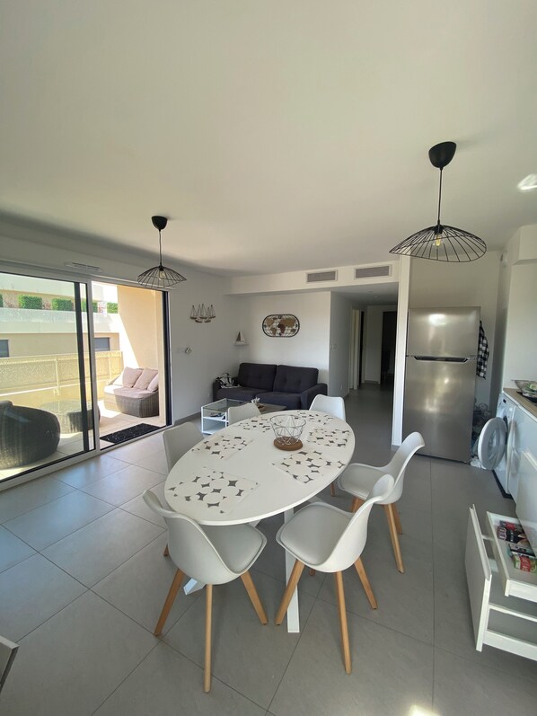 A modern kitchen with a dining area, featuring a large round table with chairs, a refrigerator, and a washing machine. - Le Refuge du Plongeur - New & Cosy Apartment (Saint-Mandrier-sur-Mer)