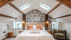 A bedroom with a stone ceiling, a bed with white and brown bedding, a round ottoman, a chair, and a small table with a lamp. - Suites at Derby House (Castletown)