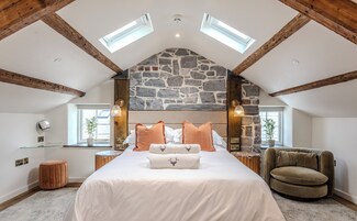 Un dormitorio con techo de piedra, una cama con ropa de cama blanca y marrón, un reposapiés redondo, una silla y una mesita con una lámpara.