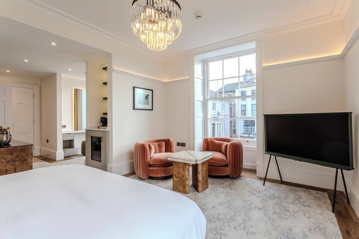 A hotel room with a large bed, a TV on a stand, two cushioned chairs, a small table, a chandelier, and a large window with a view of buildings.