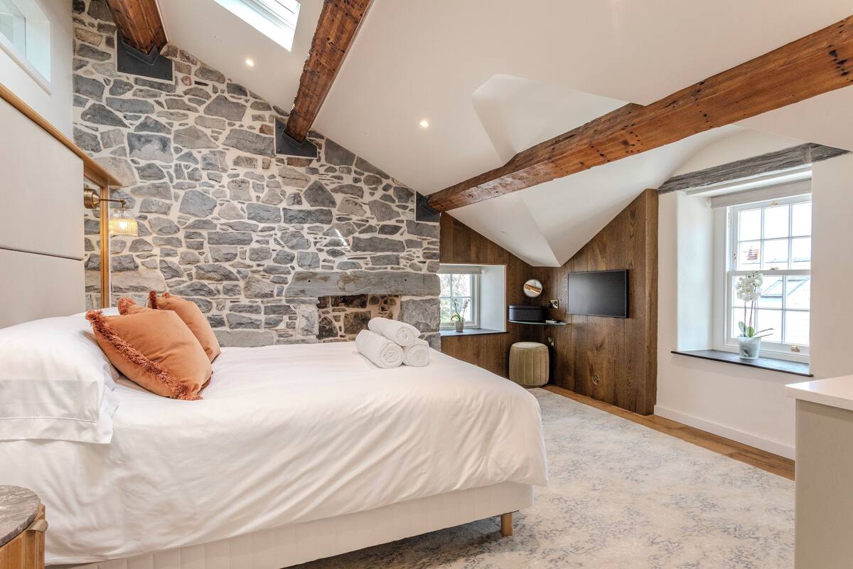 A bedroom with a stone wall, wooden beams, a bed with white bedding, a flat-screen TV, and a window with a view.